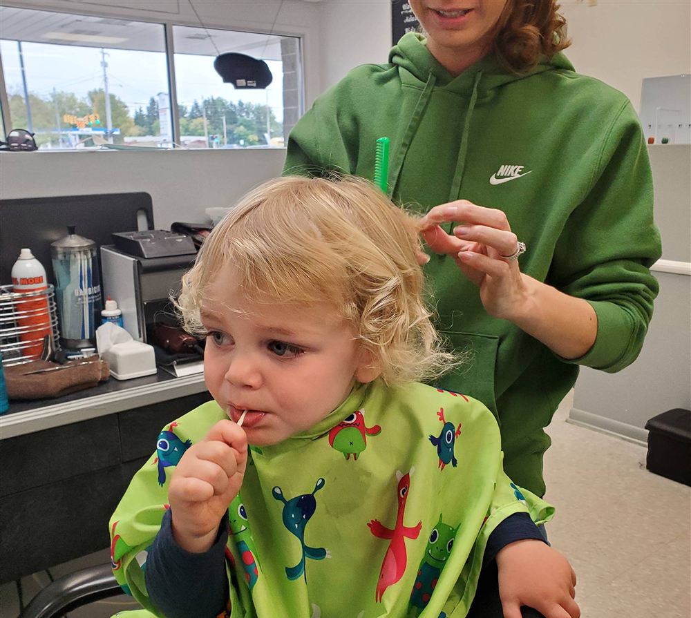 April trimming a toddler's blond curls while he eats a lollipop